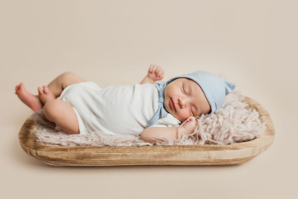 Sleeping newborn baby wearing white and blue clothing rests peacefully in a rustic wooden bowl.