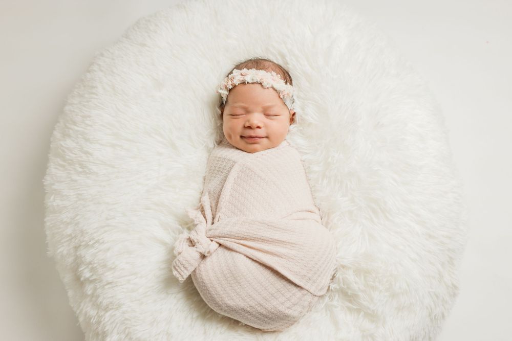 Newborn baby wrapped in soft white fabric sleeping peacefully on fluffy white surface with delicate headband.