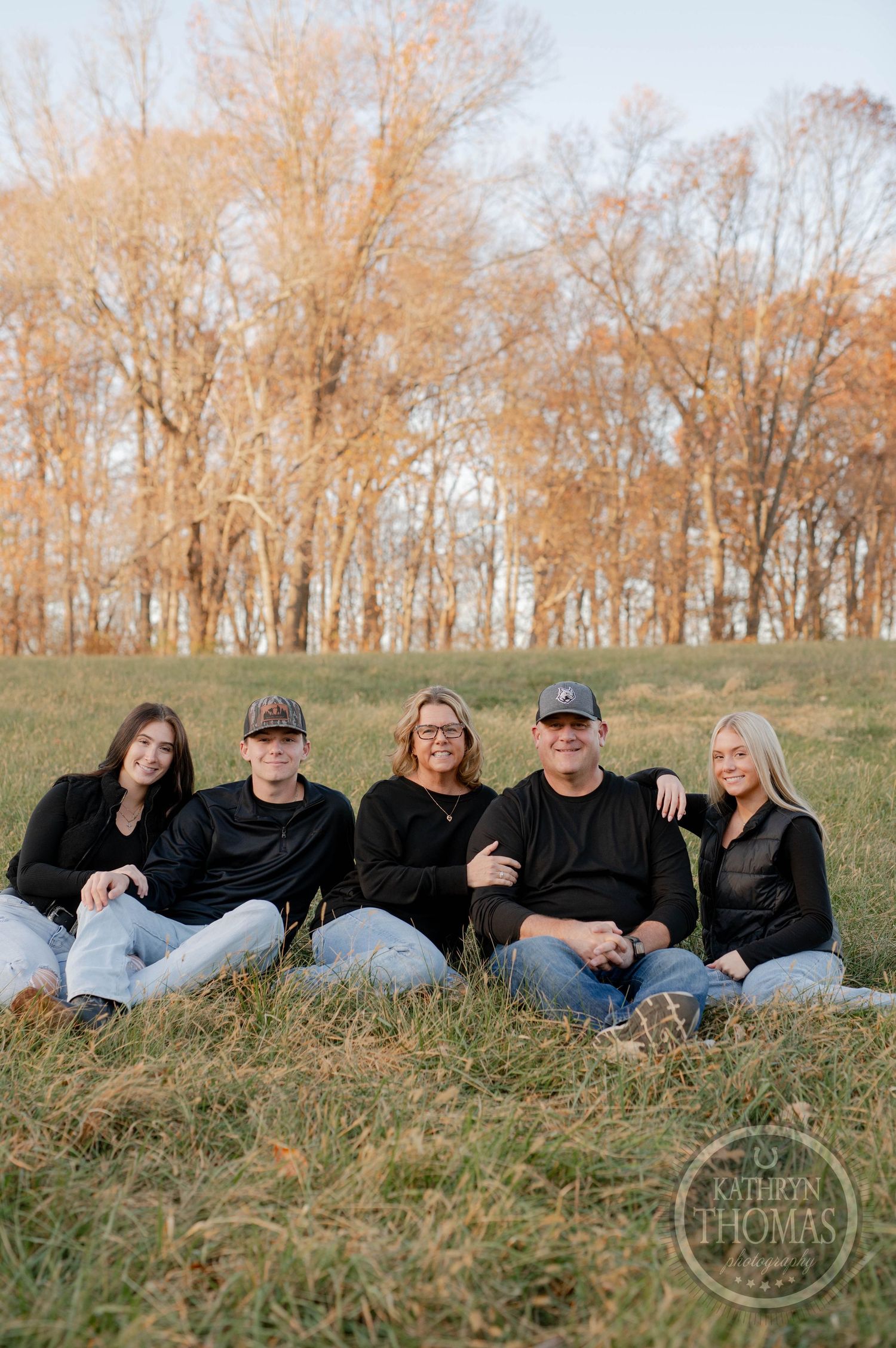 Family sitting together on grassy slope with fall trees in background during outdoor photo session.