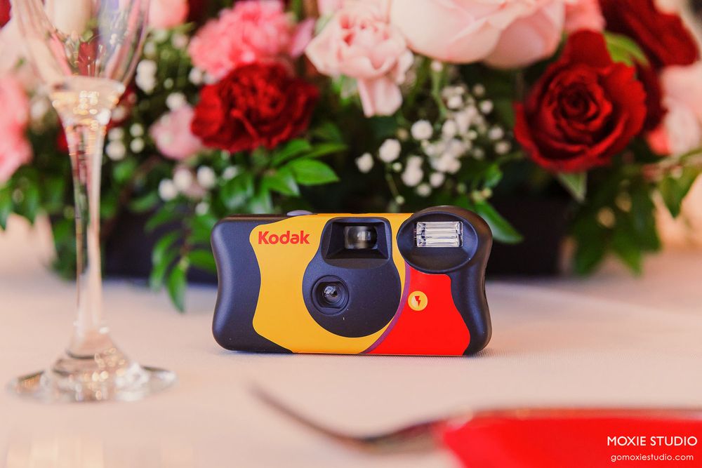 Disposable film camera sits among pink and red roses with white baby's breath flowers on a decorated table setting.