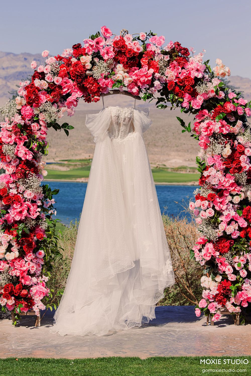 Wedding arch decorated with vibrant pink and red flowers against a lakeside mountain backdrop.