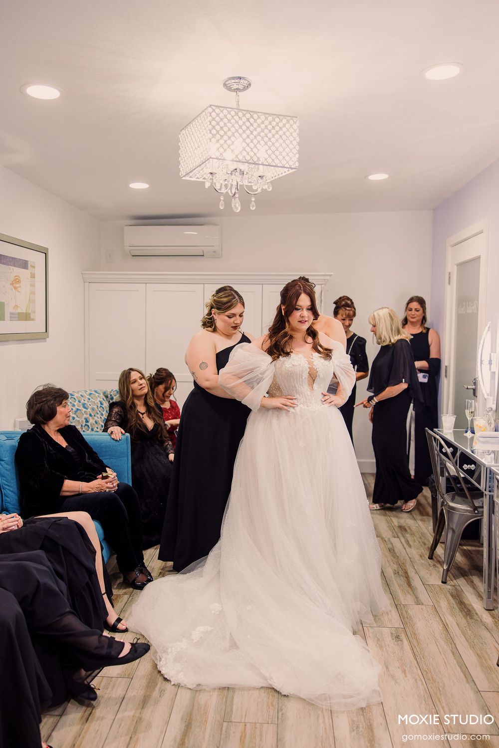 Wedding preparation scene in a bright room with people gathered around a flowing white bridal gown.