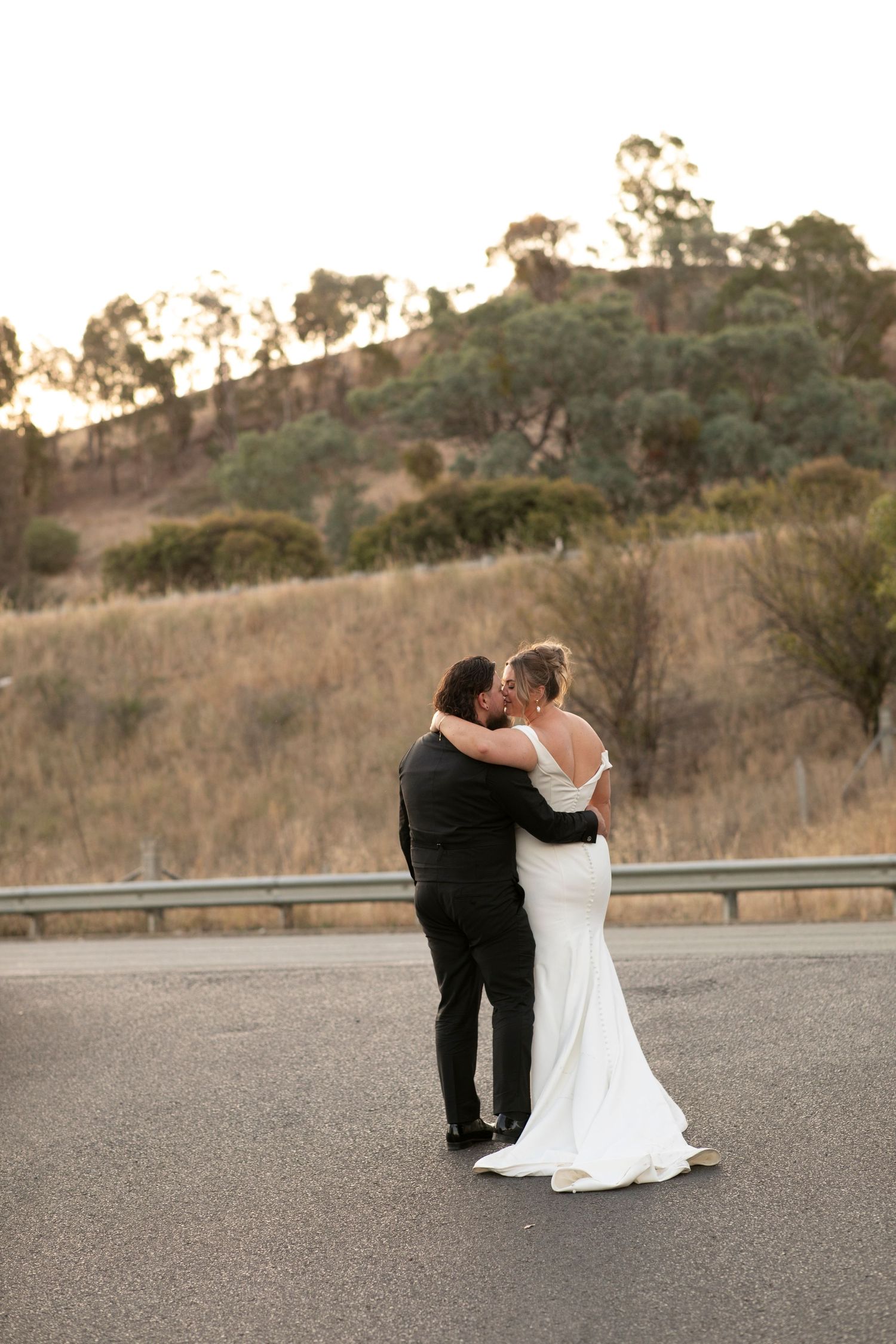 A wedding couple embraces on a rural road at sunset with rolling hills in the background.