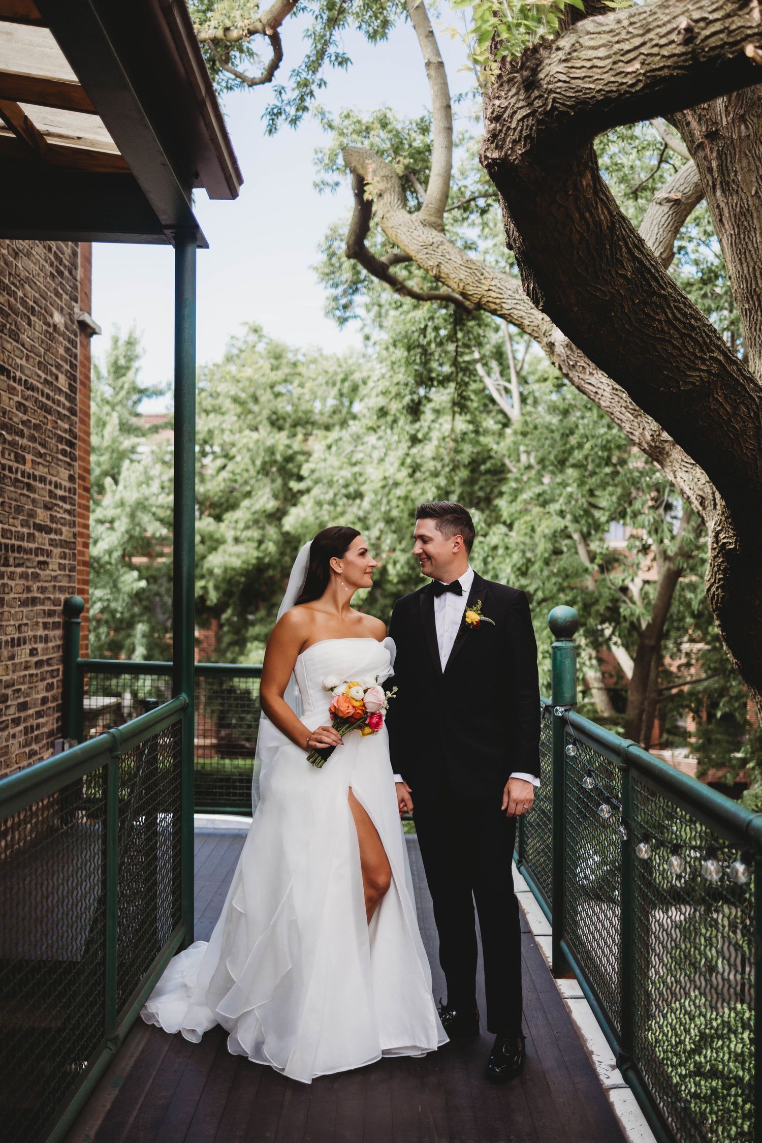 Elegant couple in wedding attire posing on outdoor deck with brick building and trees in background.