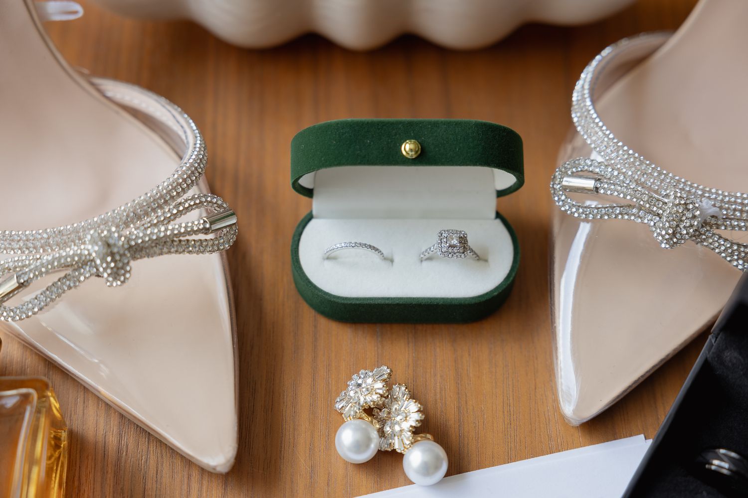 Wedding accessories including silver pointed heels, pearl earrings, and wedding rings in a green velvet box on wooden surface.