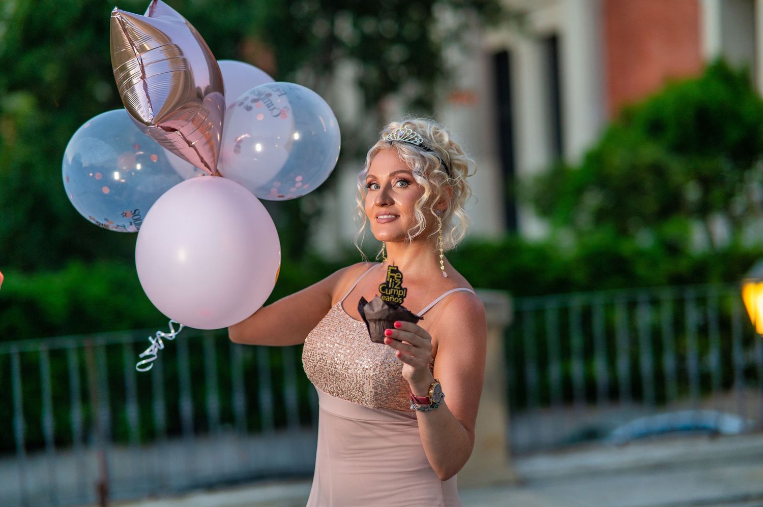 Elegant figure in pink dress holds pastel balloons and a beverage while standing outdoors at dusk.