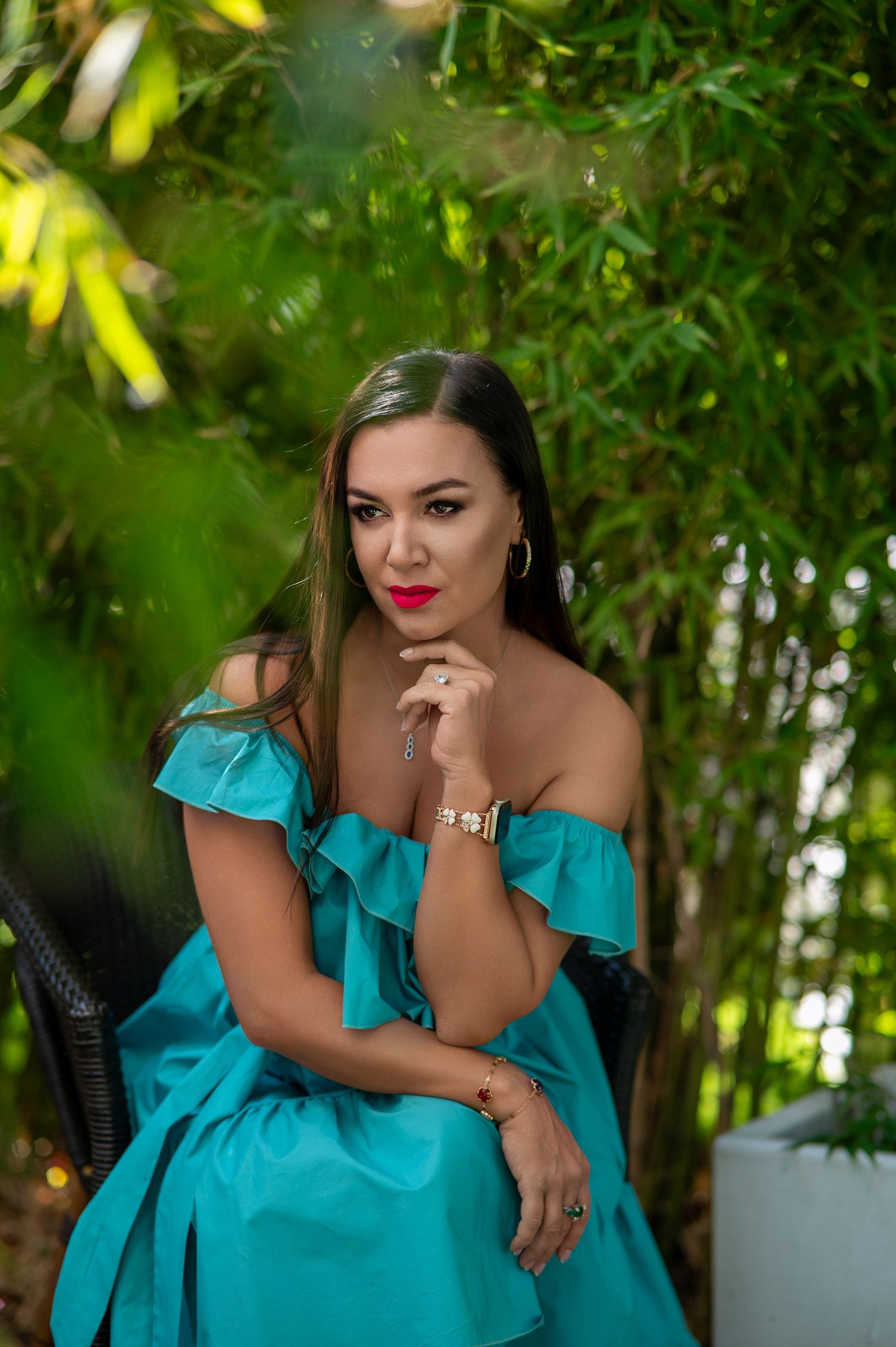 A woman in a turquoise off-shoulder dress poses thoughtfully in a garden setting with bamboo plants in the background.