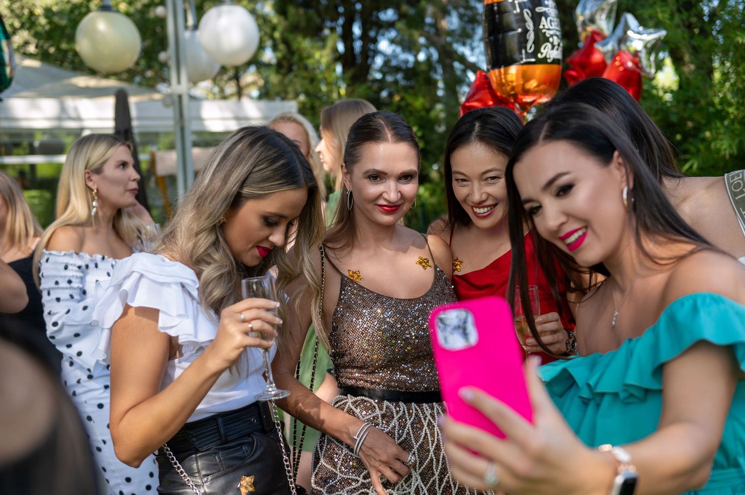 A group of elegantly dressed party guests gather at an outdoor event to look at a pink phone.