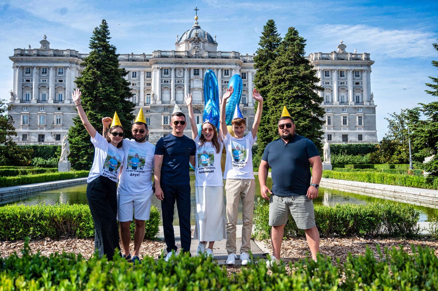A group poses cheerfully in front of the Royal Palace of Madrid on a sunny summer day with blue skies.