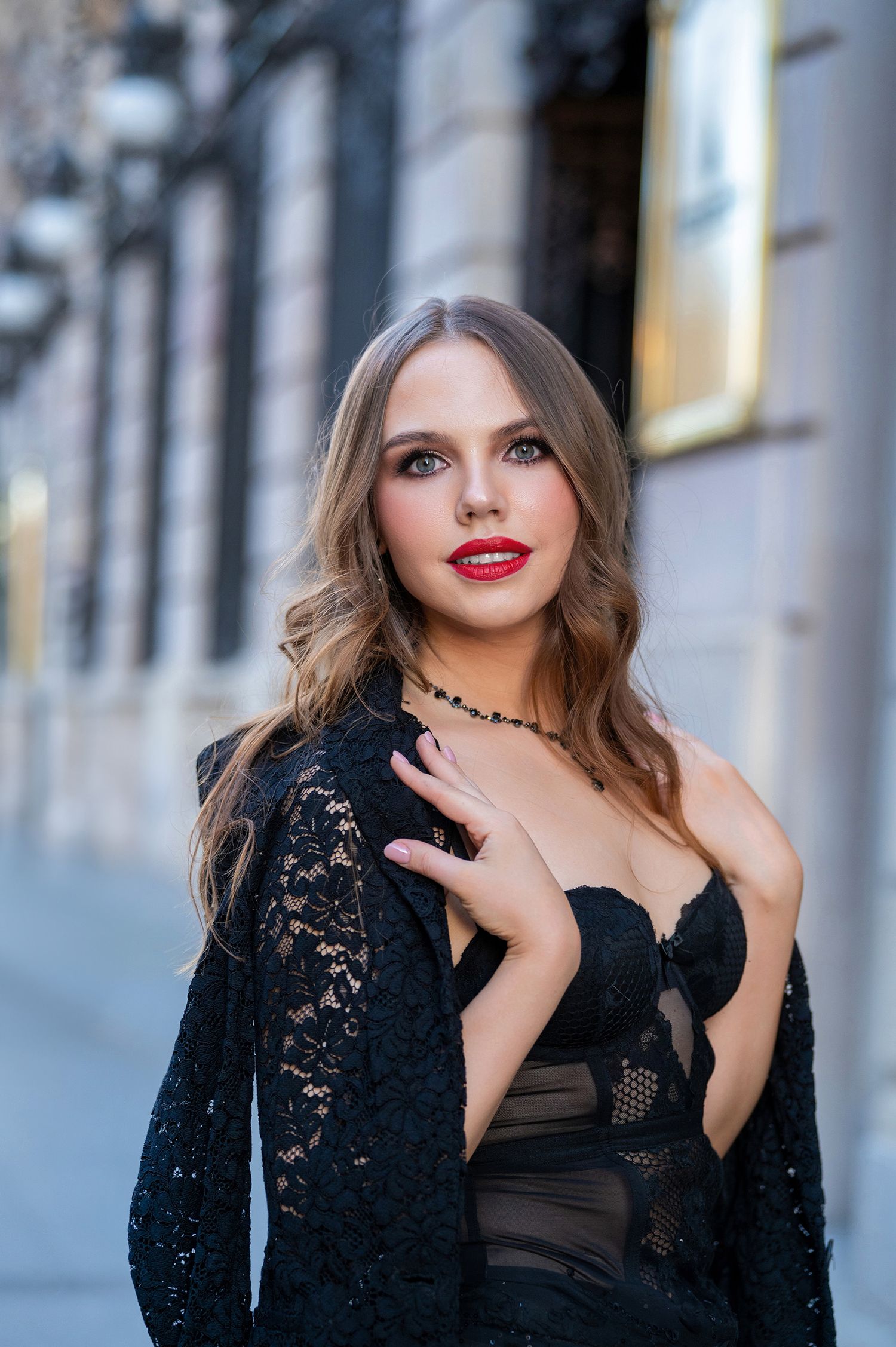 Woman in black lace jacket and lingerie with red lipstick posing elegantly on urban street.