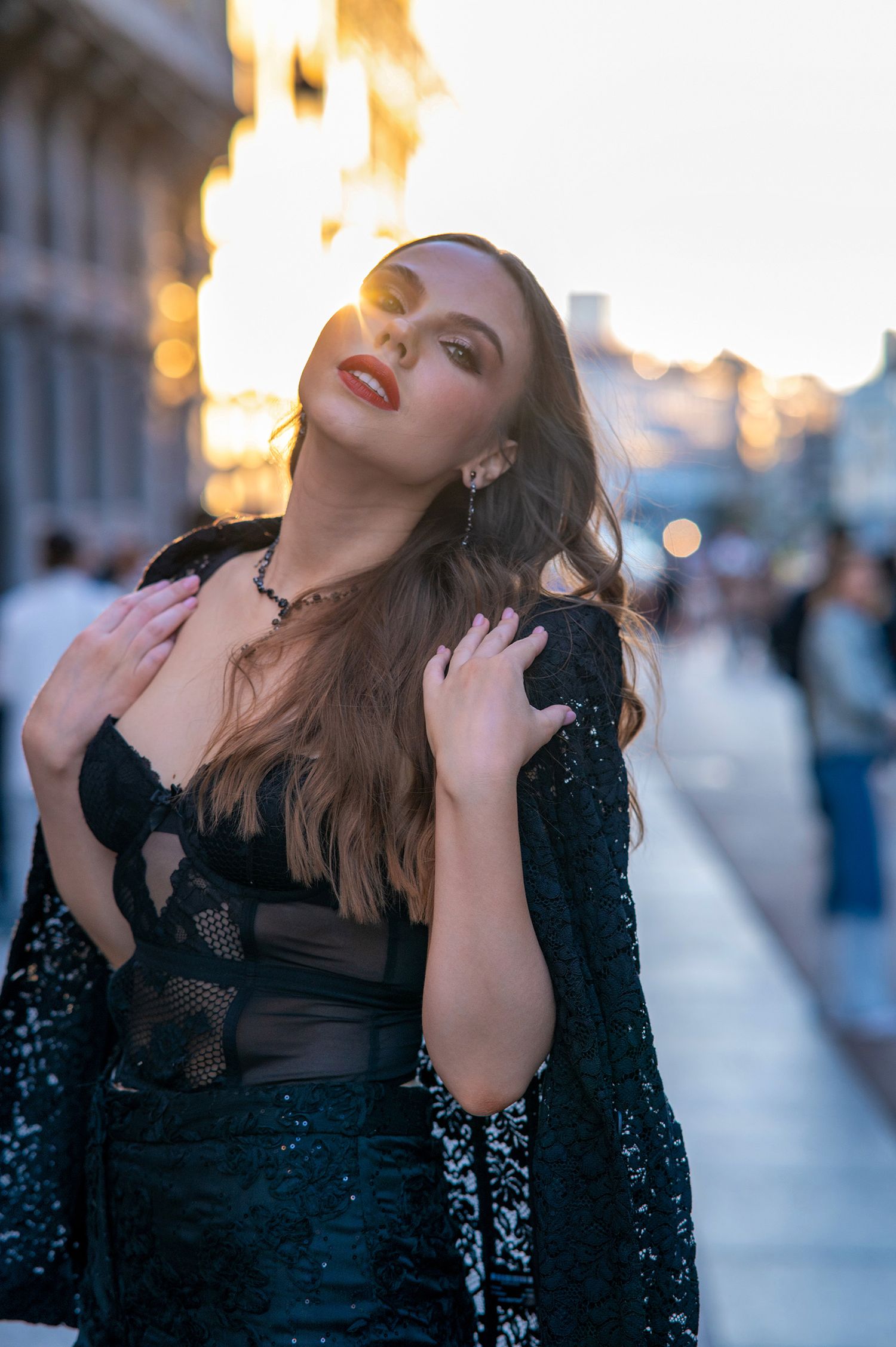 Woman in elegant black sequined gown poses on city street at sunset with glowing bokeh lights in background.