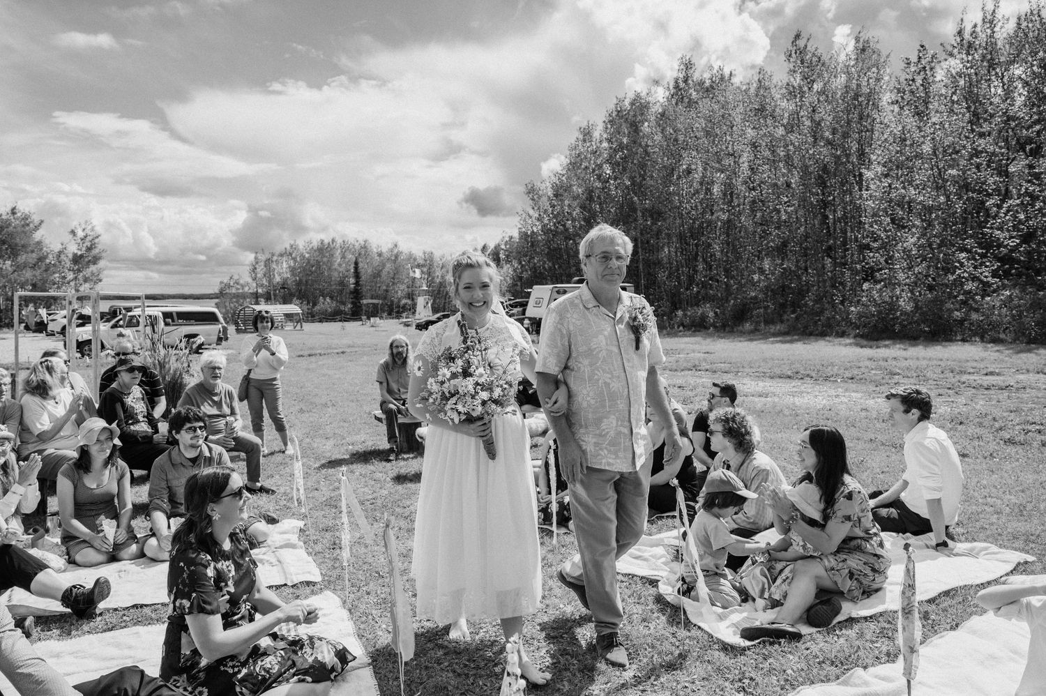 Outdoor wedding ceremony with guests seated on wooden benches in a sunlit field.