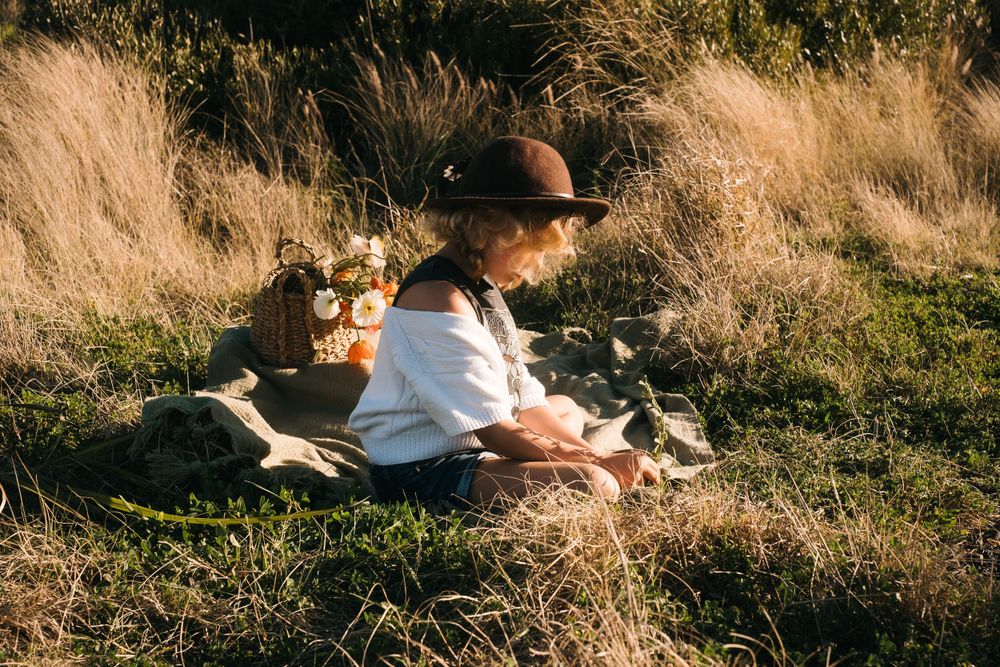 Person sits in a whimsical picnic setting with tall golden grass and vintage accessories in warm sunlight.