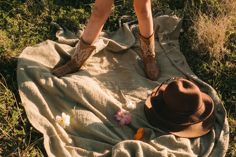 Brown cowboy boots and hat arranged on neutral blanket in sunlit outdoor setting.