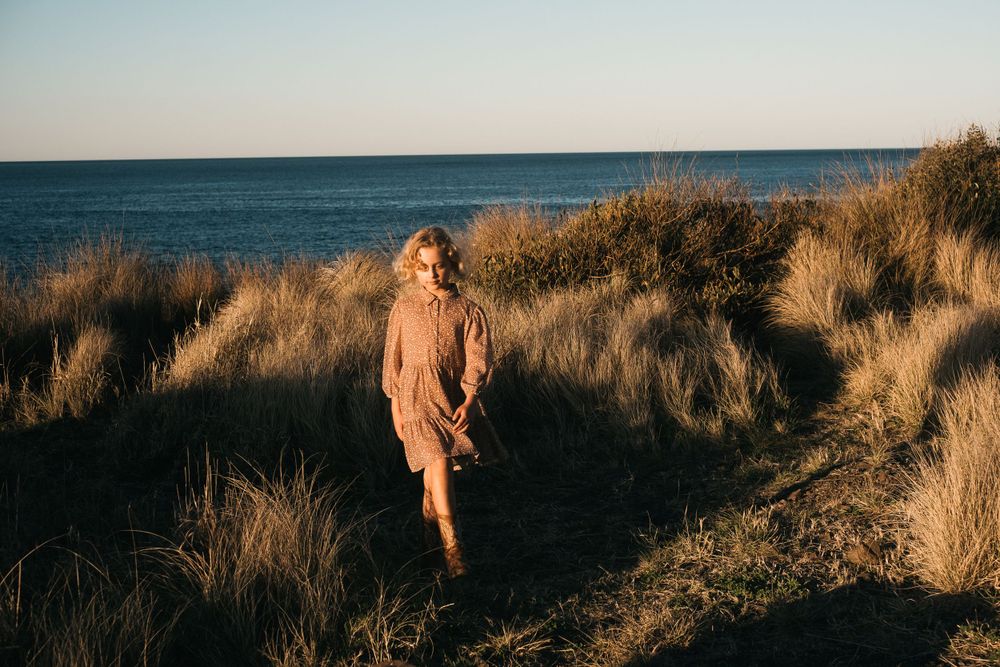 Person walking through tall coastal grasses at sunset near the ocean.