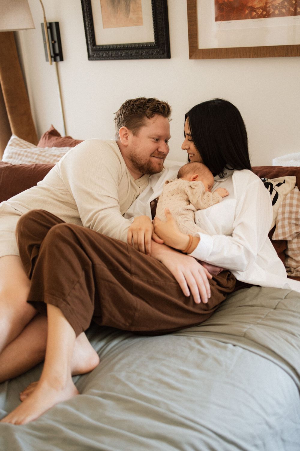Happy couple relaxes on gray bed with newborn baby in casual intimate home setting.