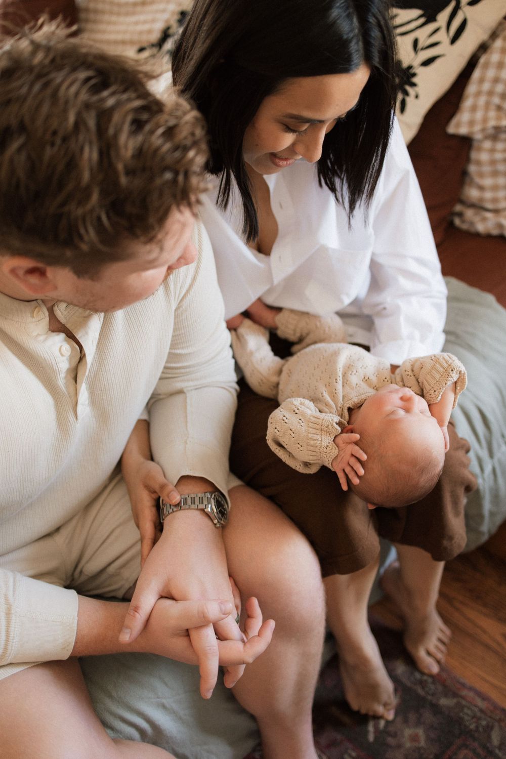 A couple sits together on a couch spending tender moments with their baby in casual white sweaters at home.