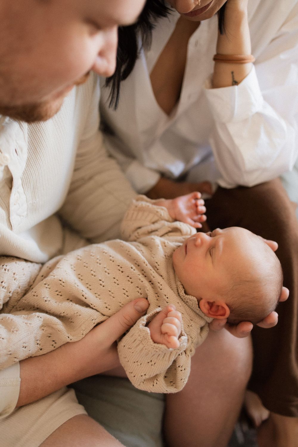 Parents gaze adoringly at their sleeping baby dressed in a cream knitted outfit.