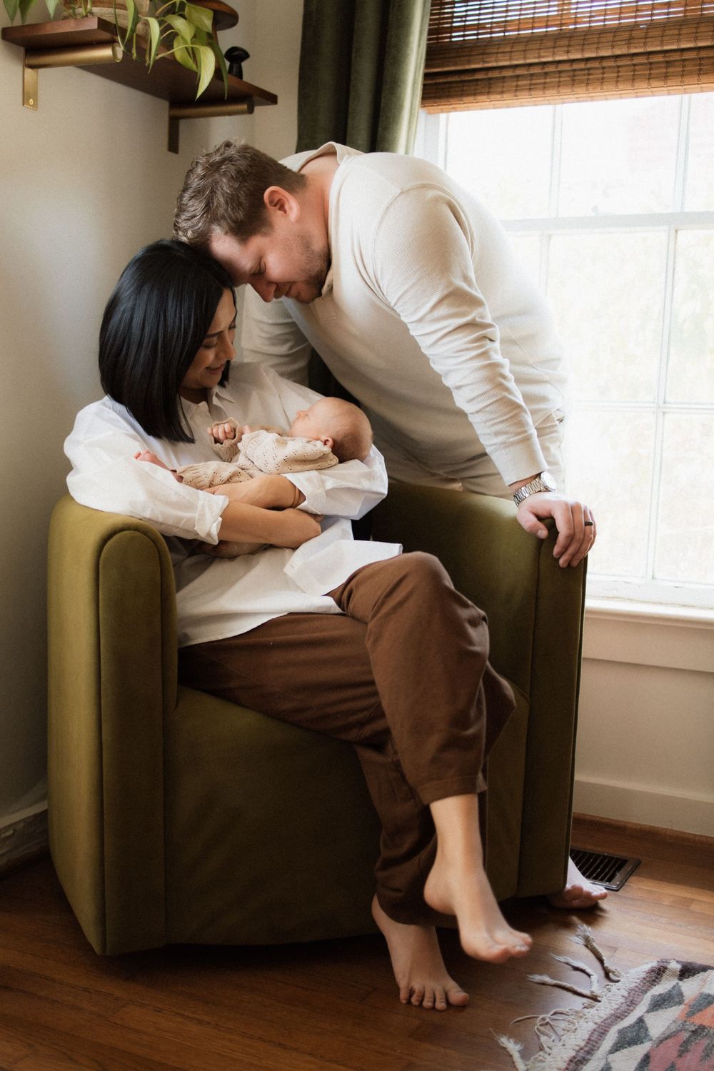 A couple shares a tender moment with their newborn baby while sitting in an olive green armchair by a window.