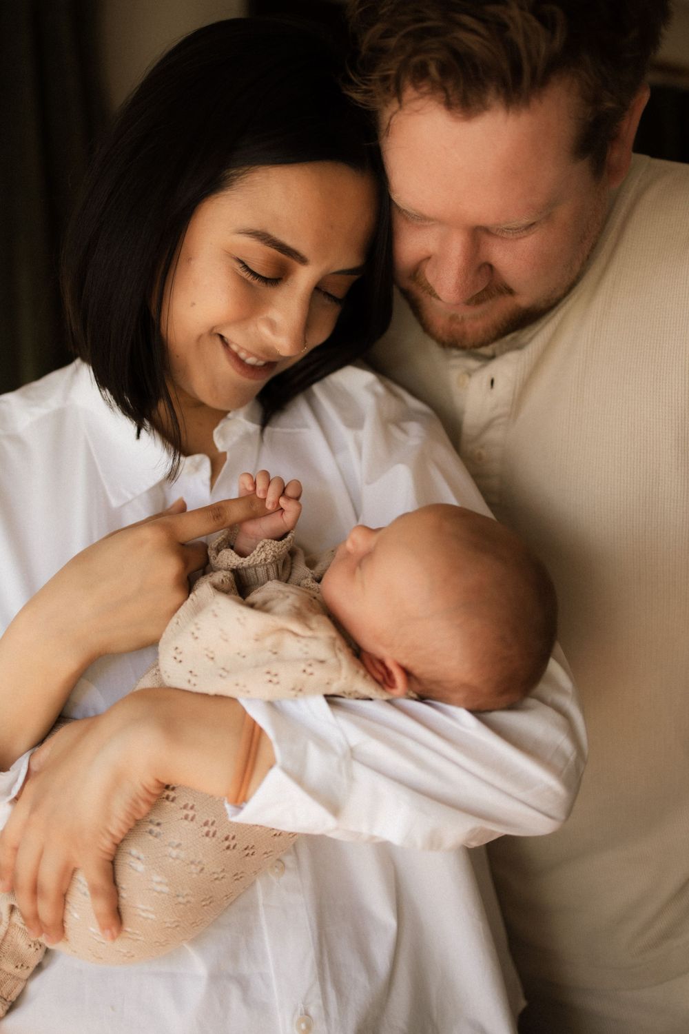 A couple shares a tender moment cuddling their newborn baby against a dark background.