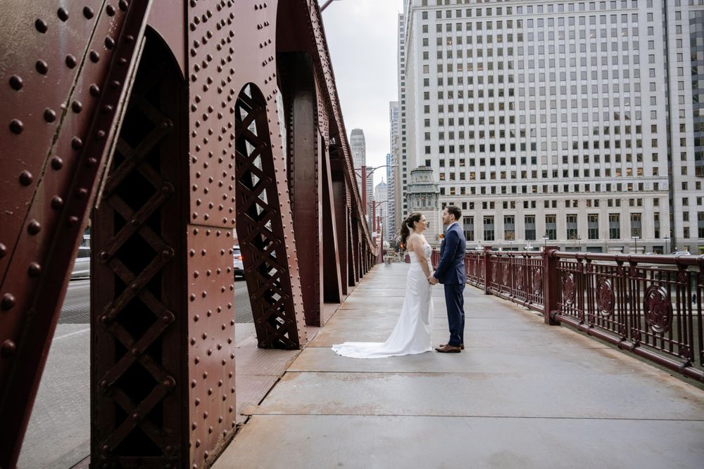 A couple stands on a historic Chicago bridge with riveted steel beams and downtown skyscrapers in the background.