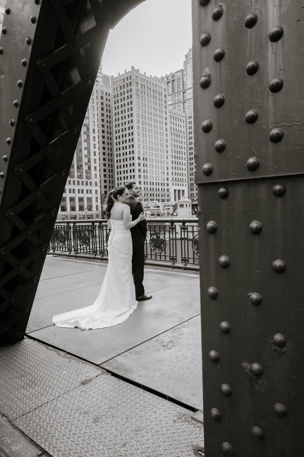 A black and white wedding photograph taken through a bridge's steel beams with a Chicago skyscraper in the background.