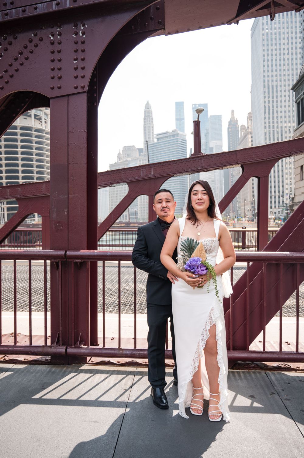 Wedding couple poses on Chicago's iconic red bridge with downtown skyline and skyscrapers in background.