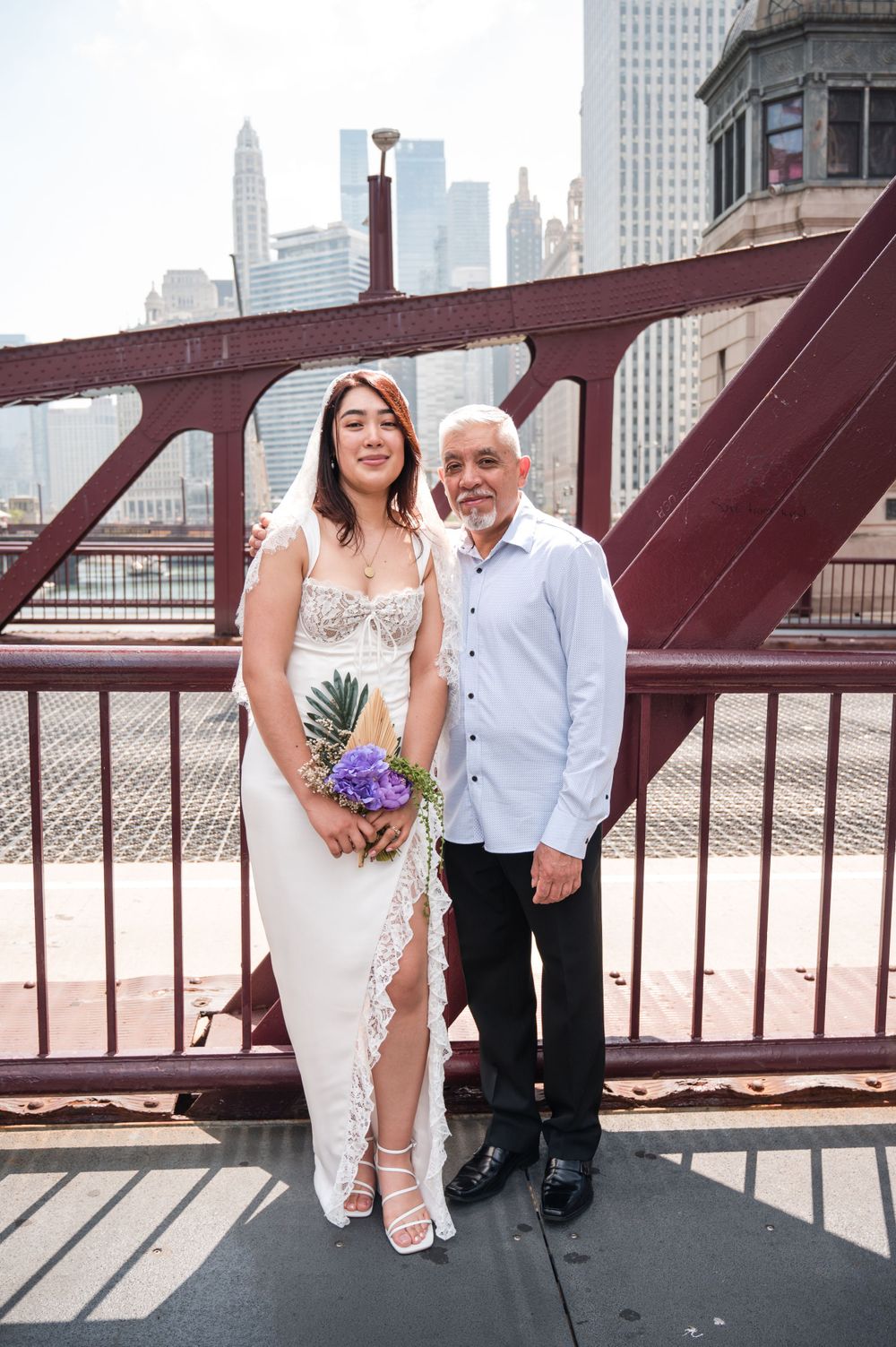 A couple poses on a Chicago bridge with the city skyline visible, the woman in a white dress holding a bouquet.