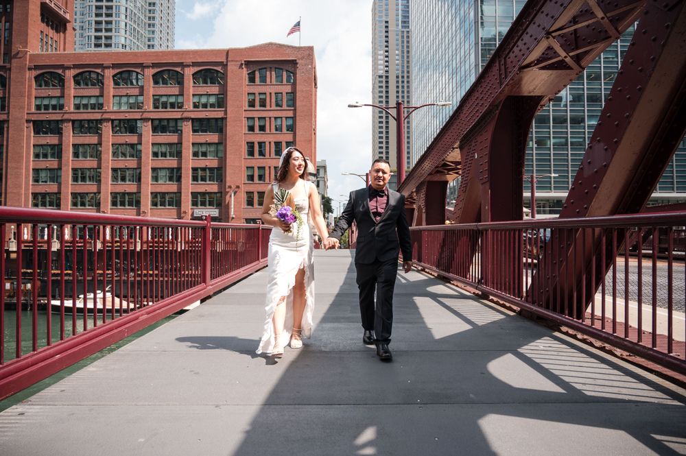 A couple walks along a red-painted bridge walkway with tall buildings visible in the background.
