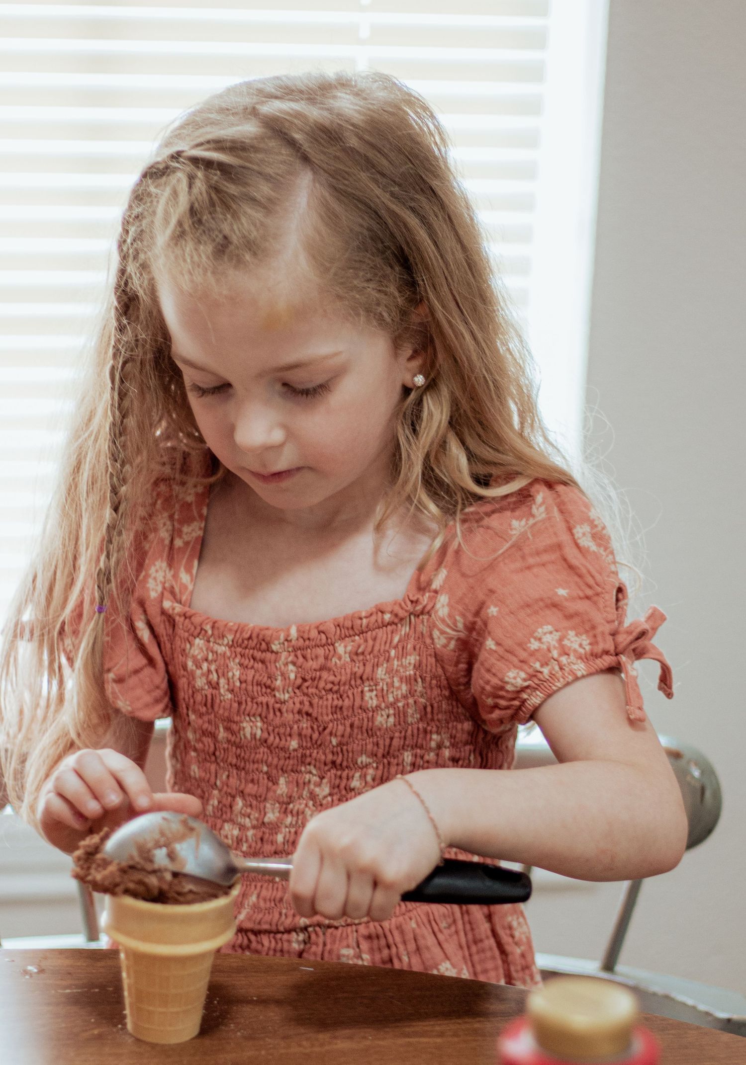 Young girl in a coral-colored smocked top scoops ice cream into a waffle cone at a wooden table.