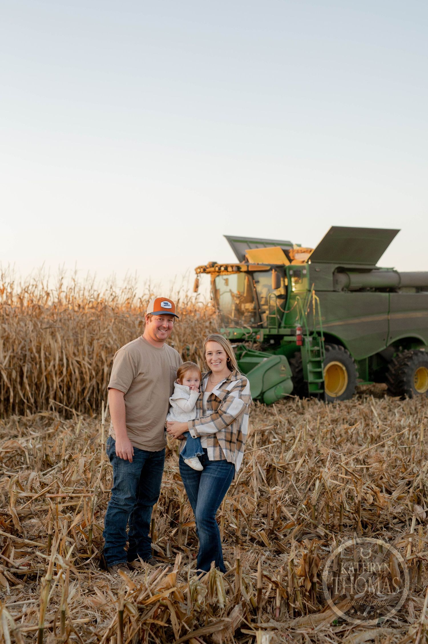 Family poses in front of green combine harvester in cornfield during harvest season.