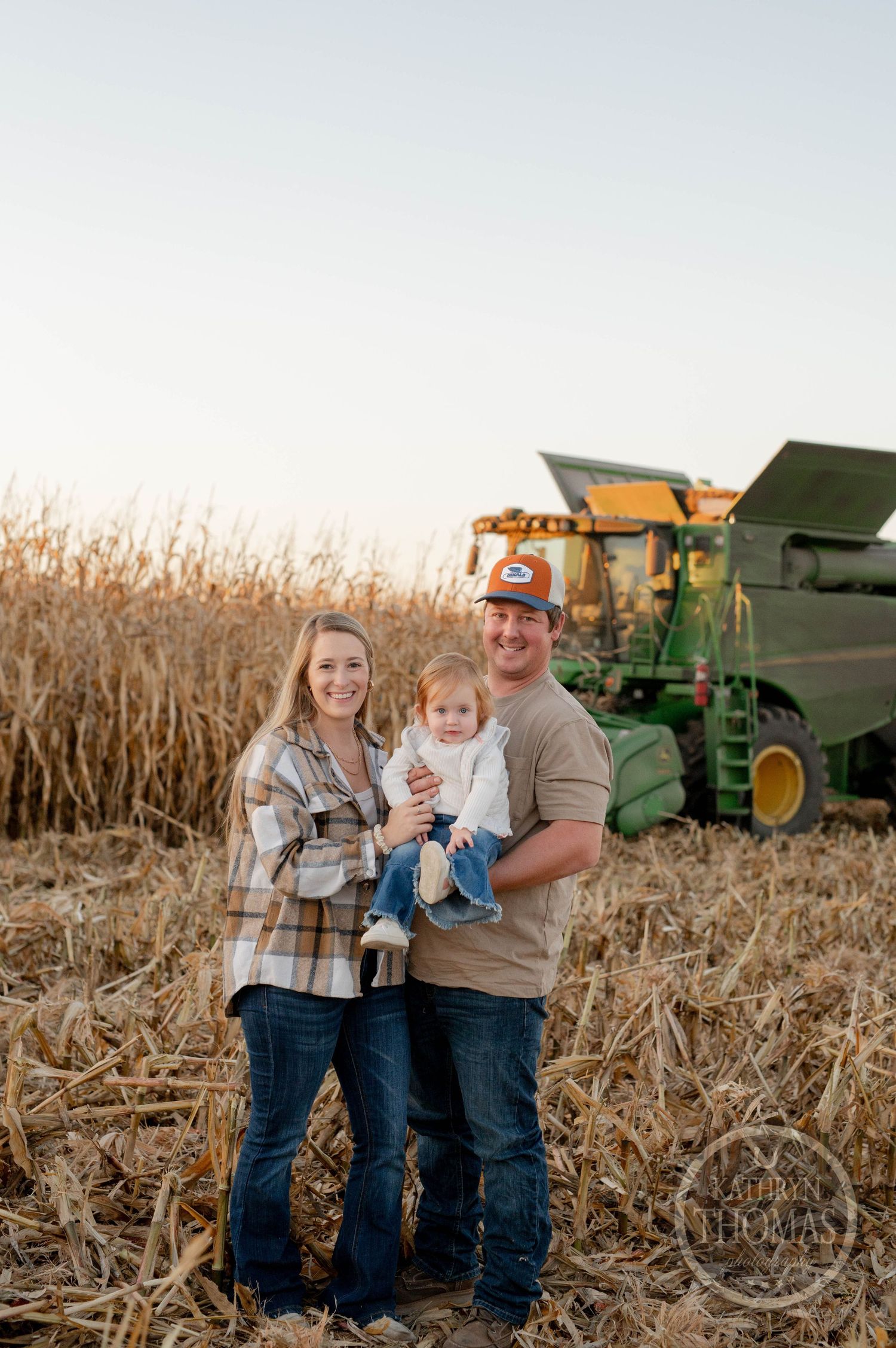 Family poses together in a cornfield during harvest with a John Deere combine in the background at sunset.