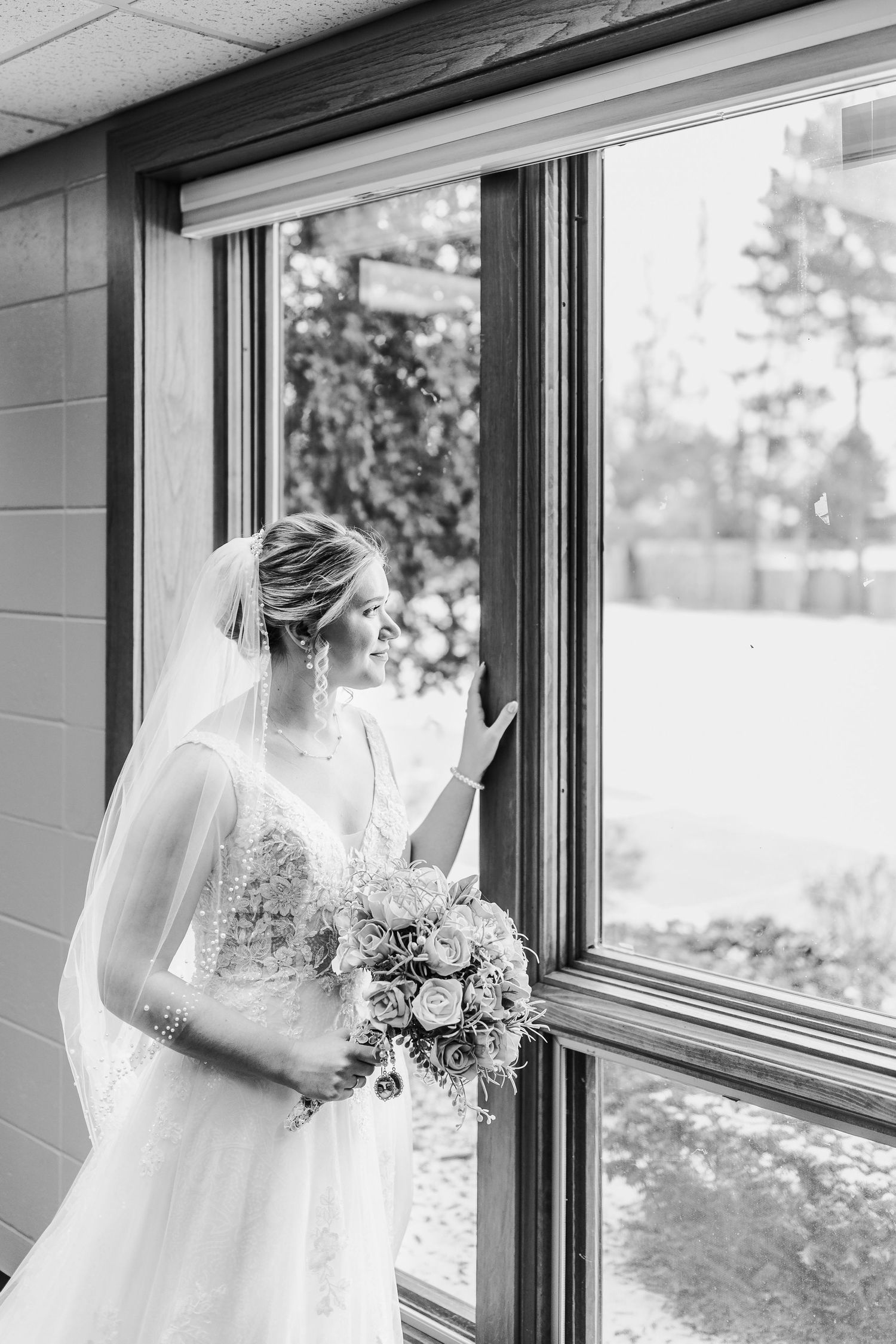 Black and white photo of bride in lace gown holding bouquet while looking out large window in contemplative moment.