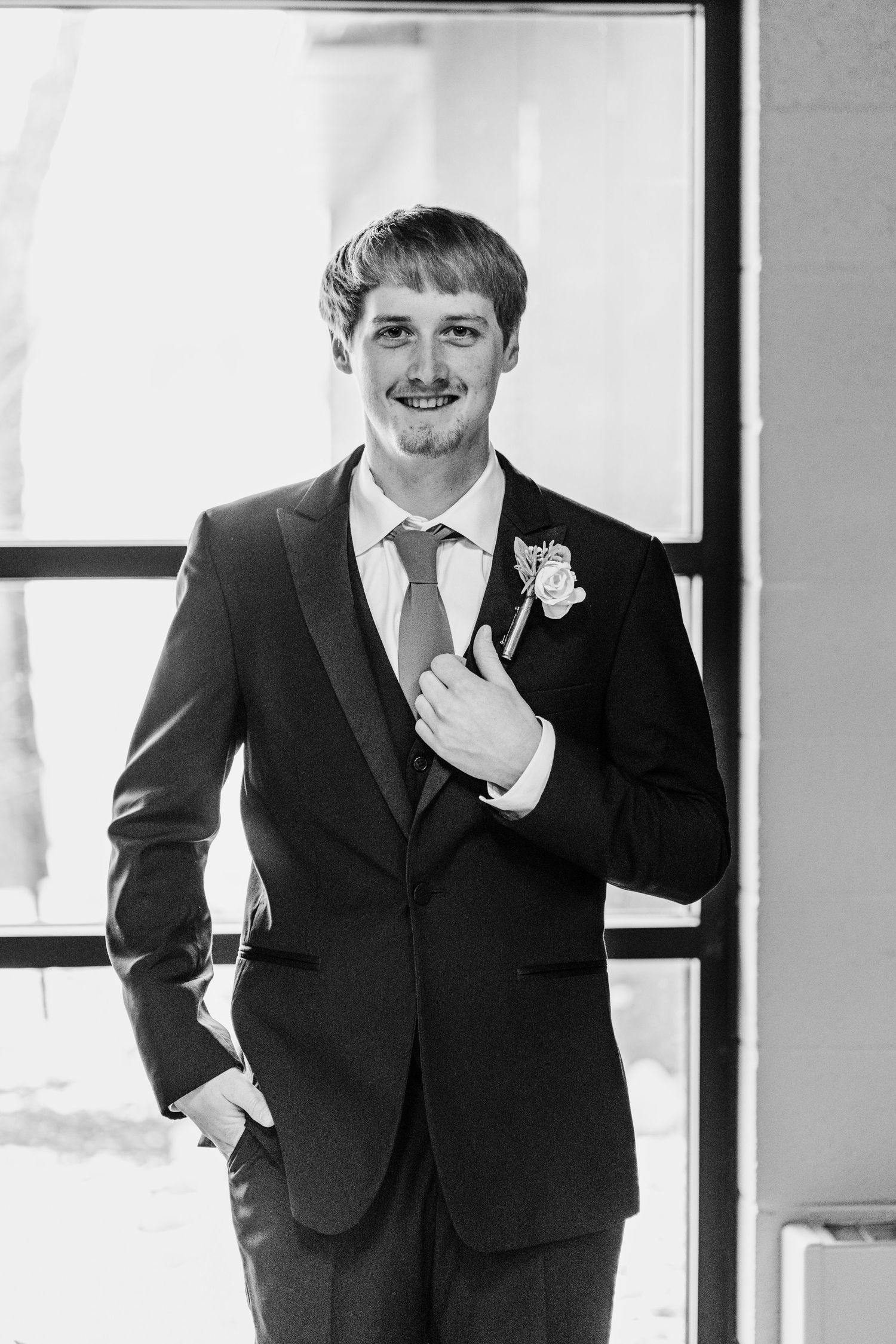 Black and white portrait of groom in formal tuxedo with boutonniere posing by a window.