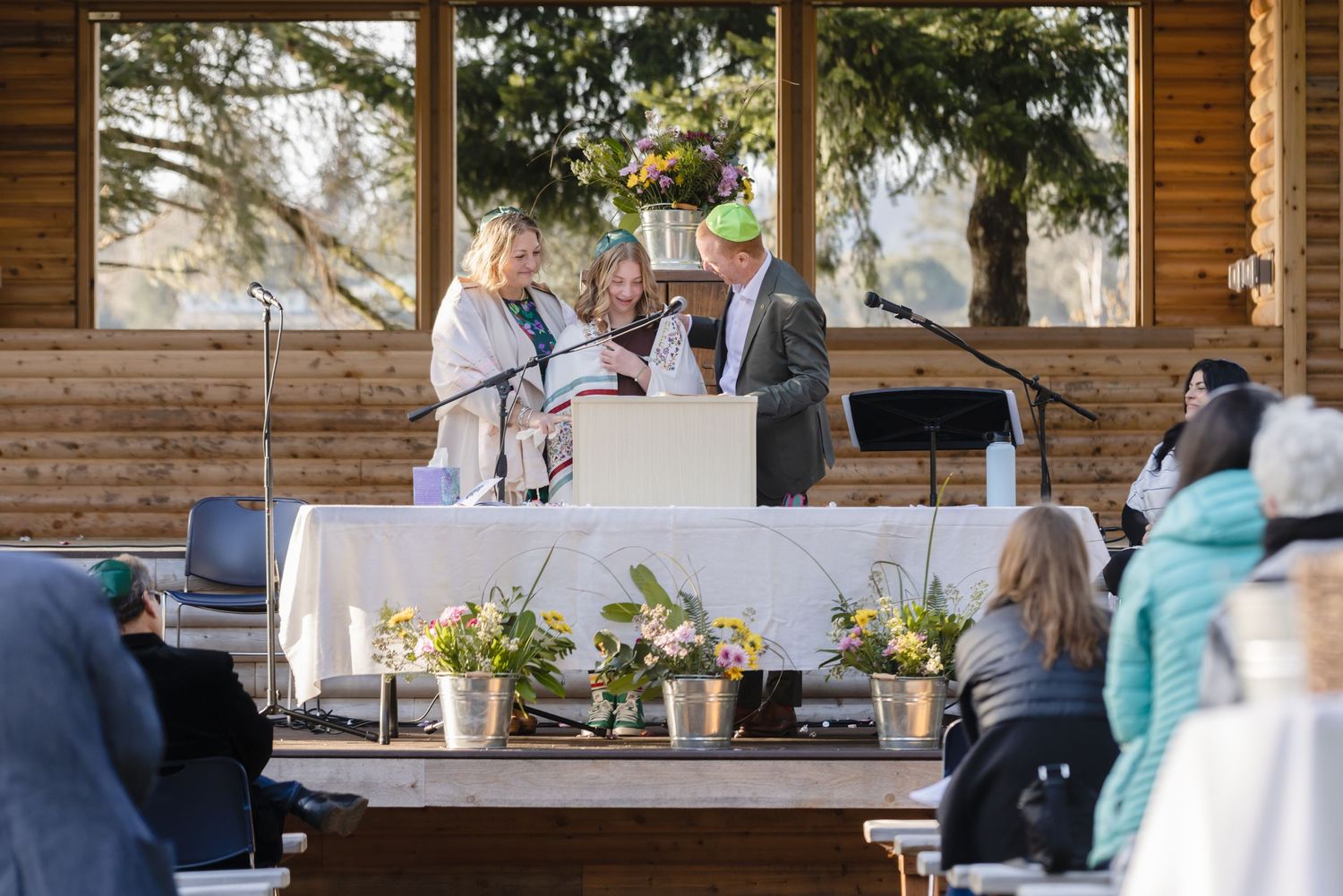 An outdoor Bat Mitzvah ceremony takes place on a rustic wooden deck with floral arrangements on the altar table.