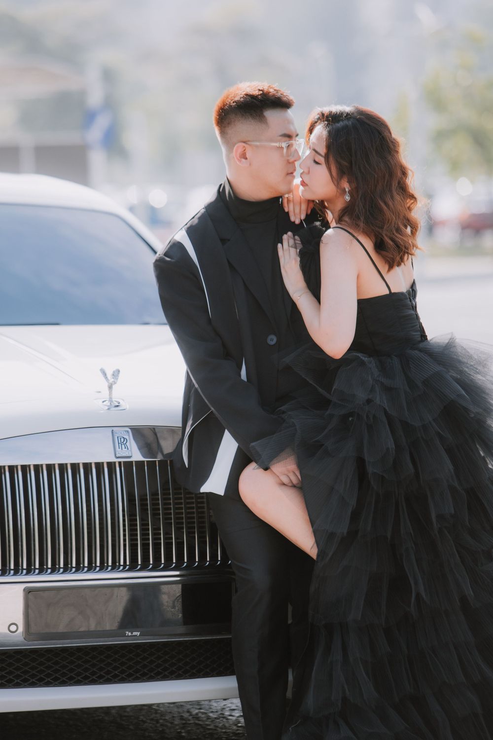 Romantic couple embracing against luxury car in matching black formal attire.