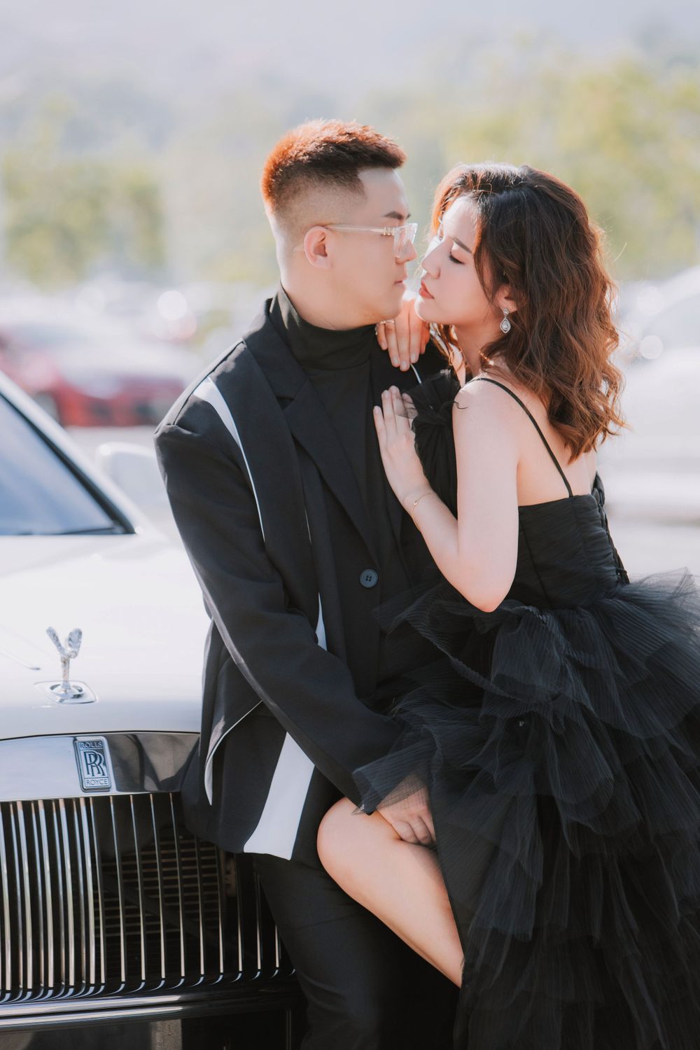 A romantic couple in formal attire pose intimately next to a luxury Rolls Royce car.