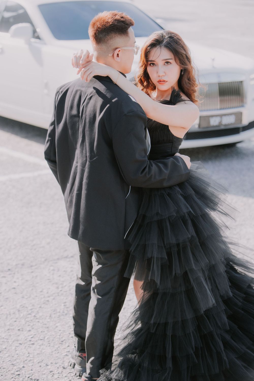 Stylish couple in formal black attire posing together near a car, with her wearing a dramatic layered tulle gown.