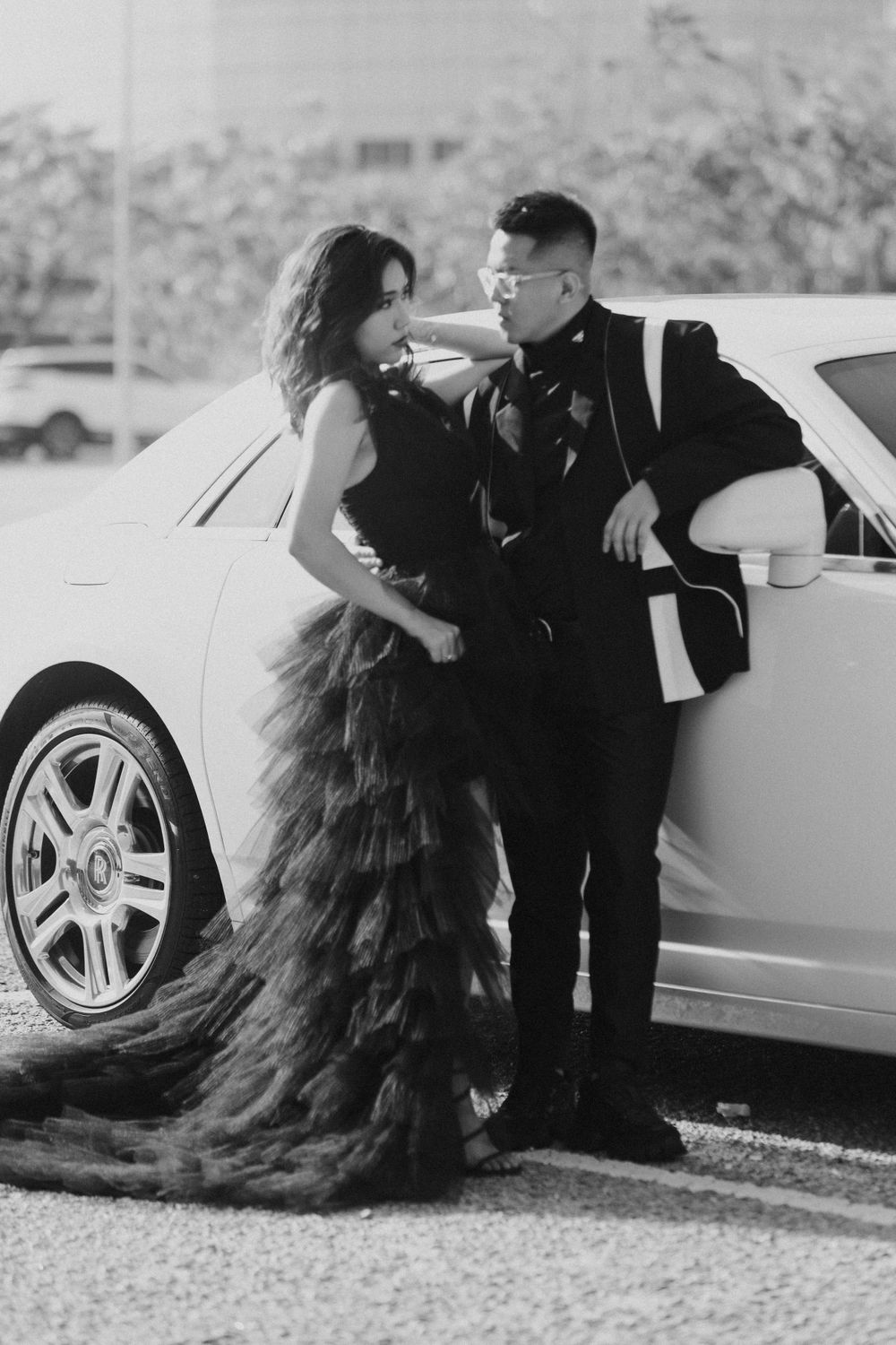 A couple poses in formal attire against a luxury car, with the woman wearing a dramatic black tulle gown.