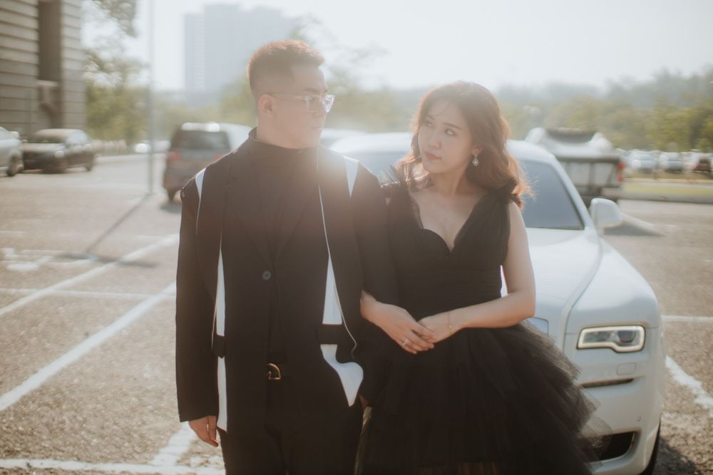 A couple in elegant black attire share a joyful moment together in a sunlit parking lot beside a luxury car.