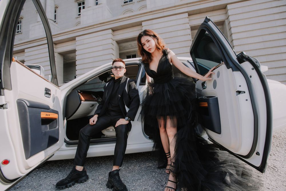 Two people with formal attire sit in a white luxury car with doors open during a stylish photoshoot.
