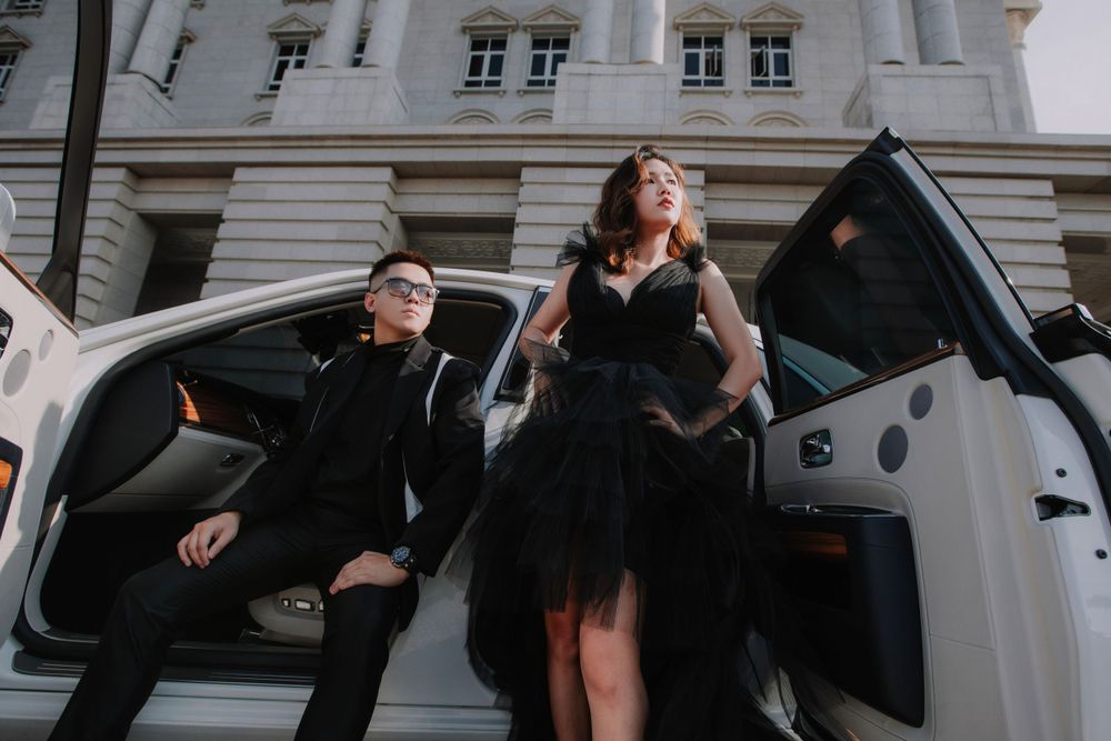 A couple in black formal attire poses dramatically by a white luxury car in front of an elegant building.