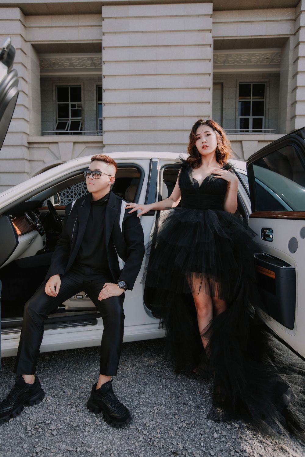 Elegantly dressed pair pose with luxury car, featuring dramatic black tulle gown and sleek suit styling.