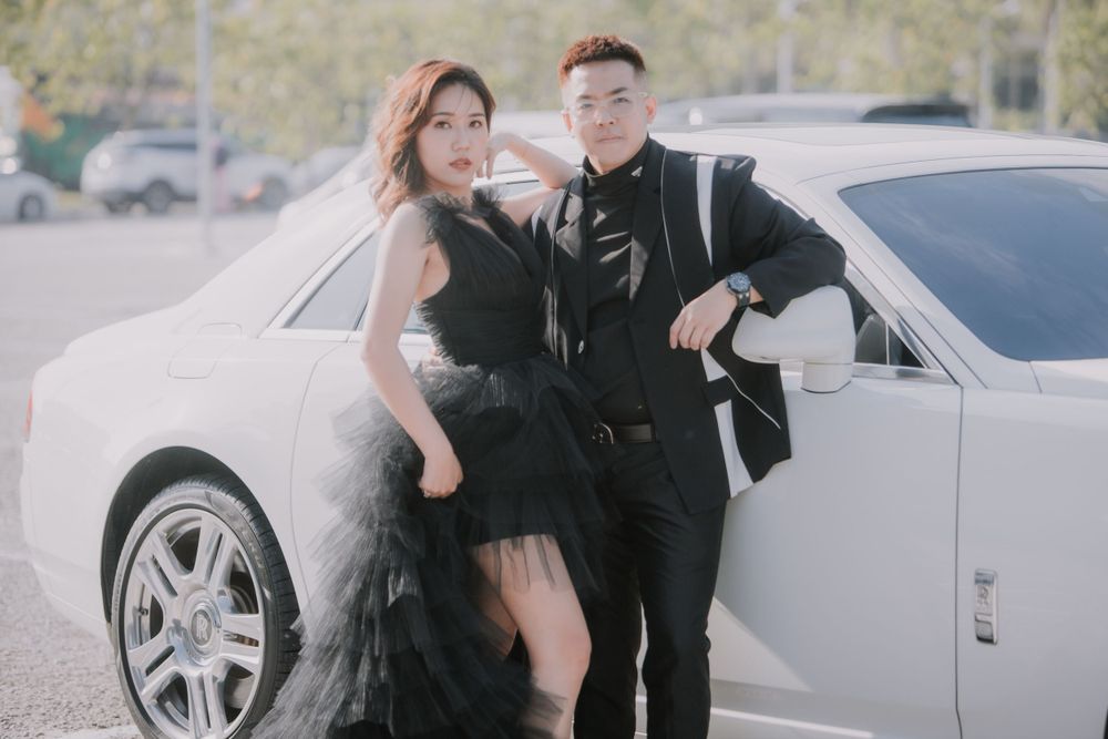 A couple poses stylishly in black outfits next to a white luxury Rolls Royce vehicle.