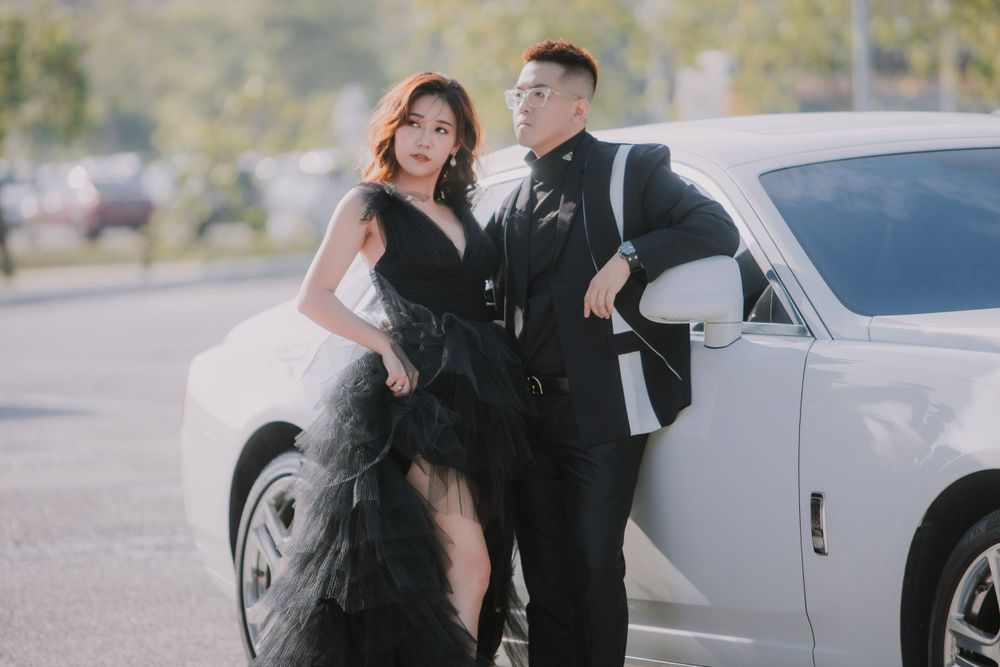 Glamorous couple in black formal wear pose against white luxury car in outdoor photoshoot.