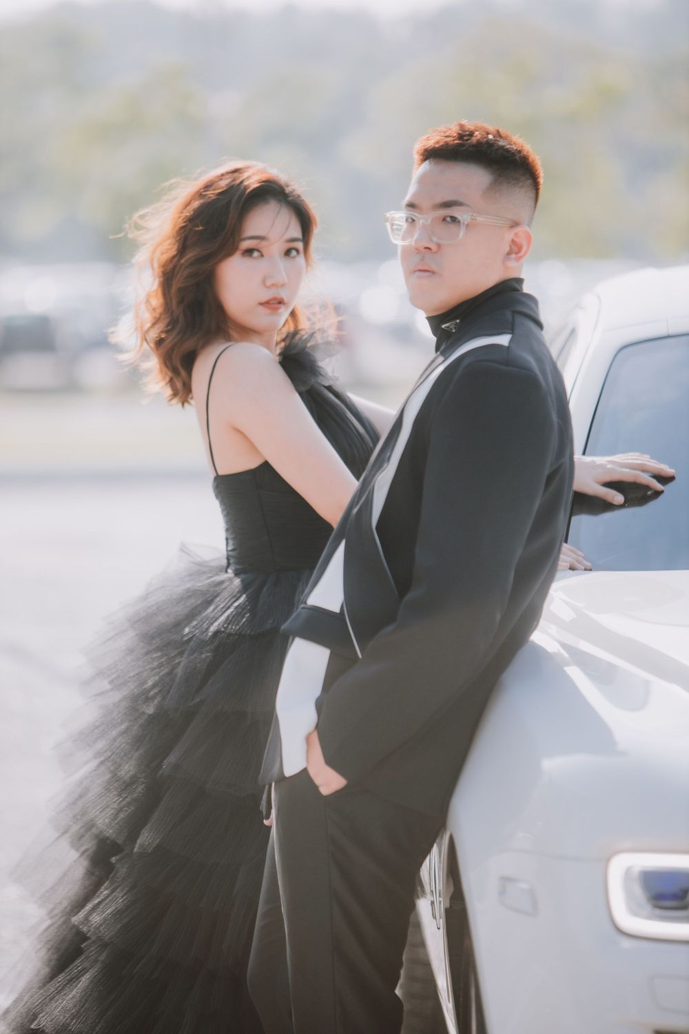 A couple in formal attire poses against a silver car, with her in a black tulle gown and him in a dark suit.