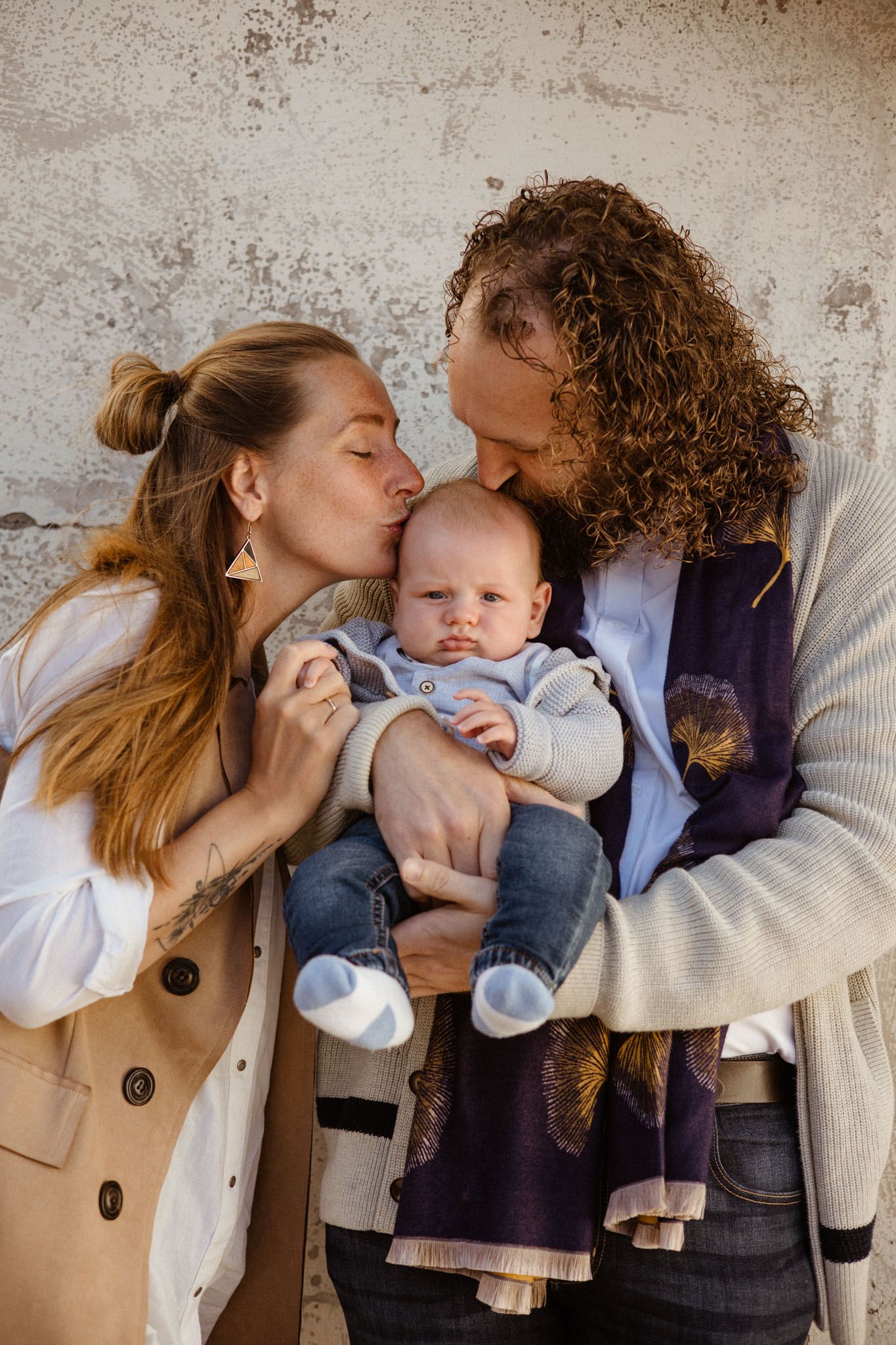 A couple sharing a tender moment with their baby outdoors against a textured wall background.