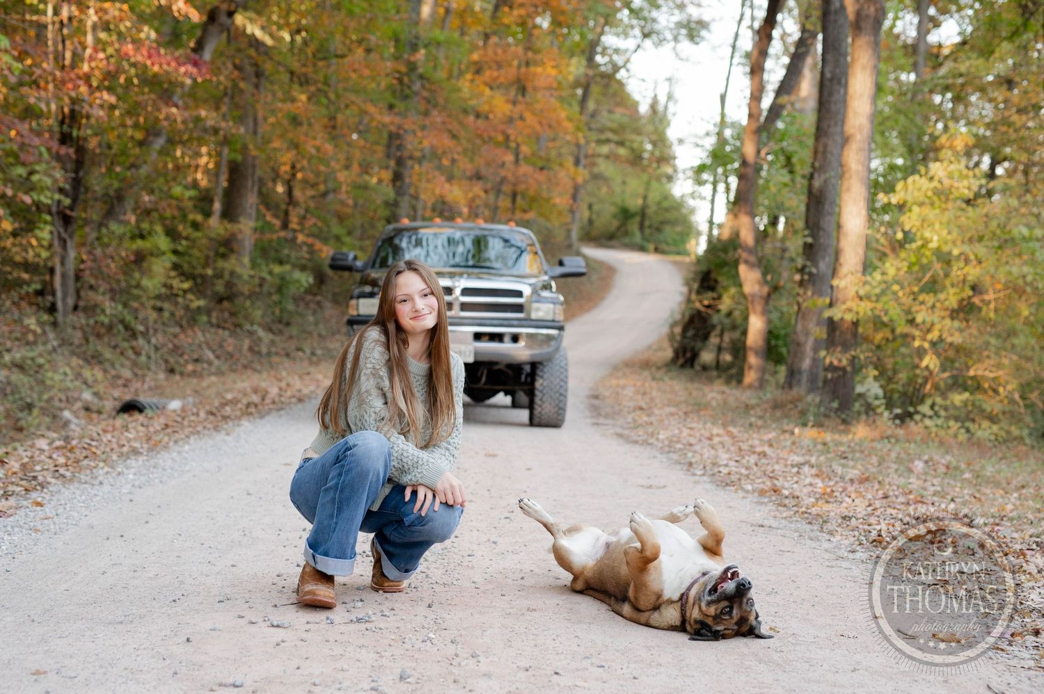 Person crouching on dirt road with playful dog rolling on ground near parked truck.