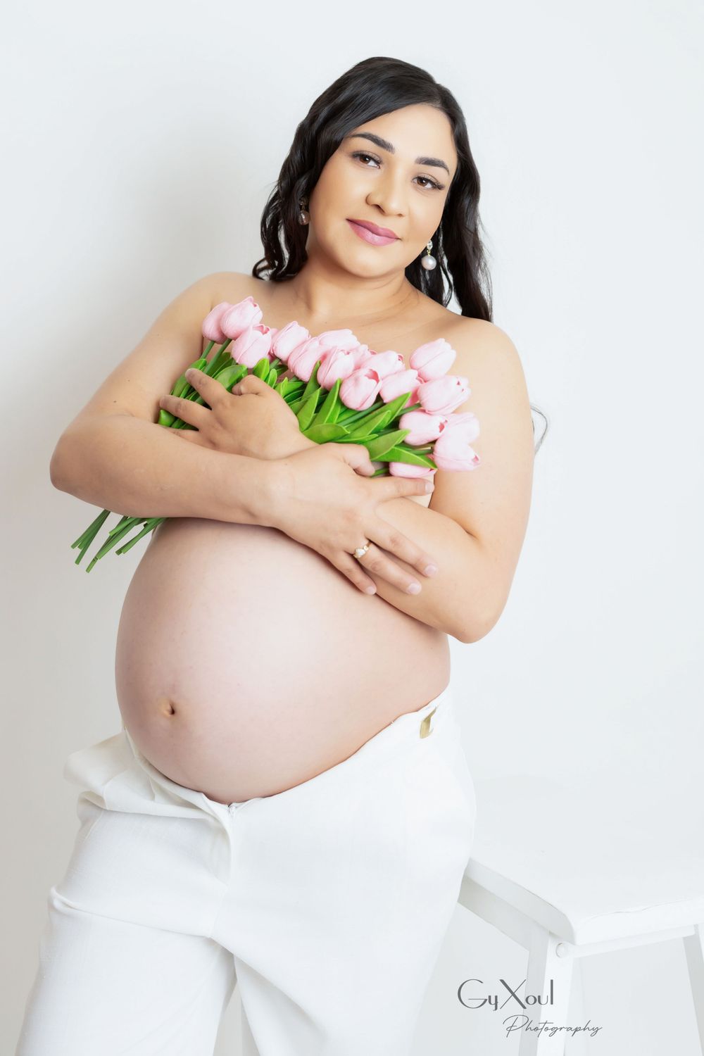 Maternity photo of woman in white clothing holding pink tulips against white background.