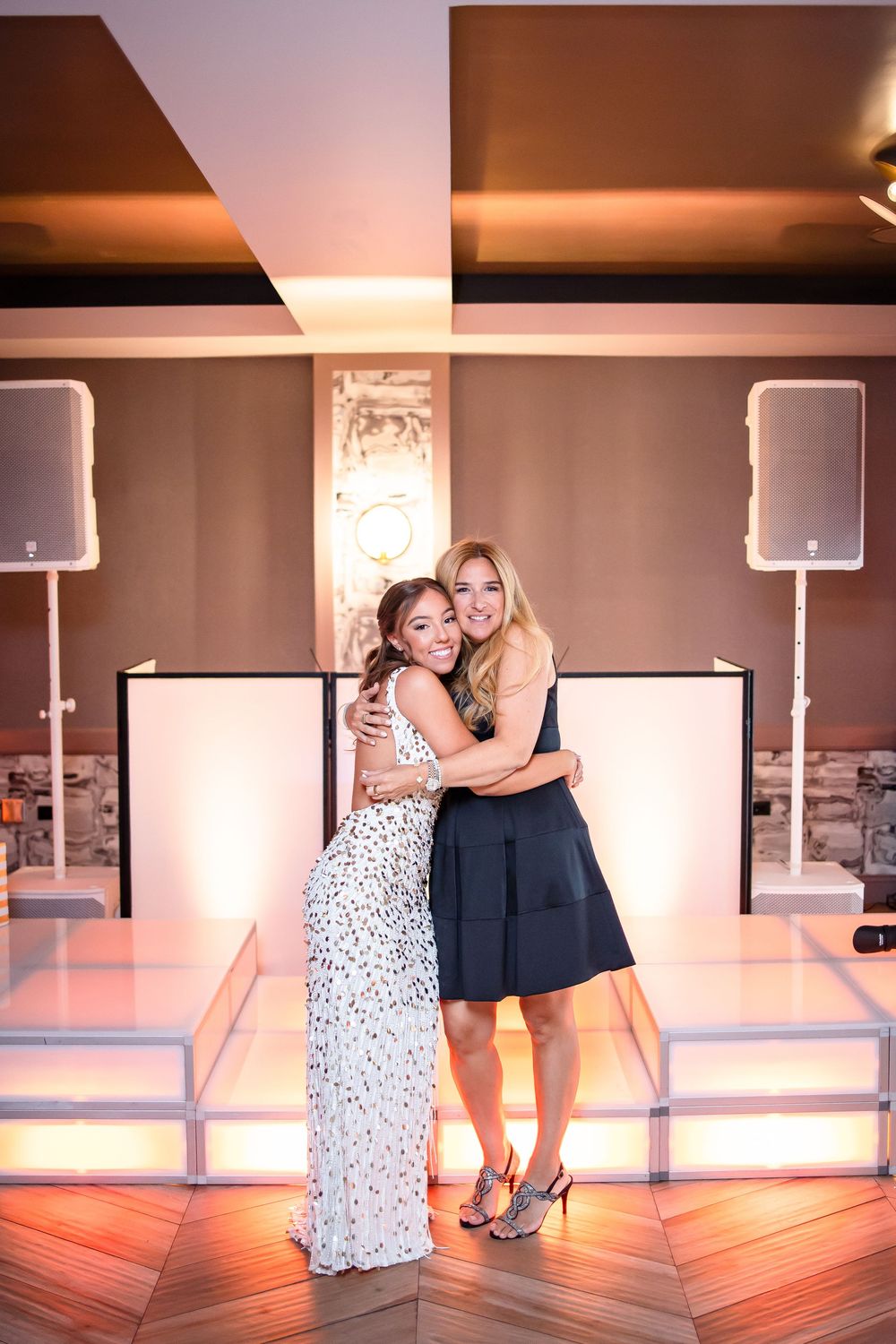 Two people in evening attire embrace on a dance floor with speakers and lighting equipment visible in the background.